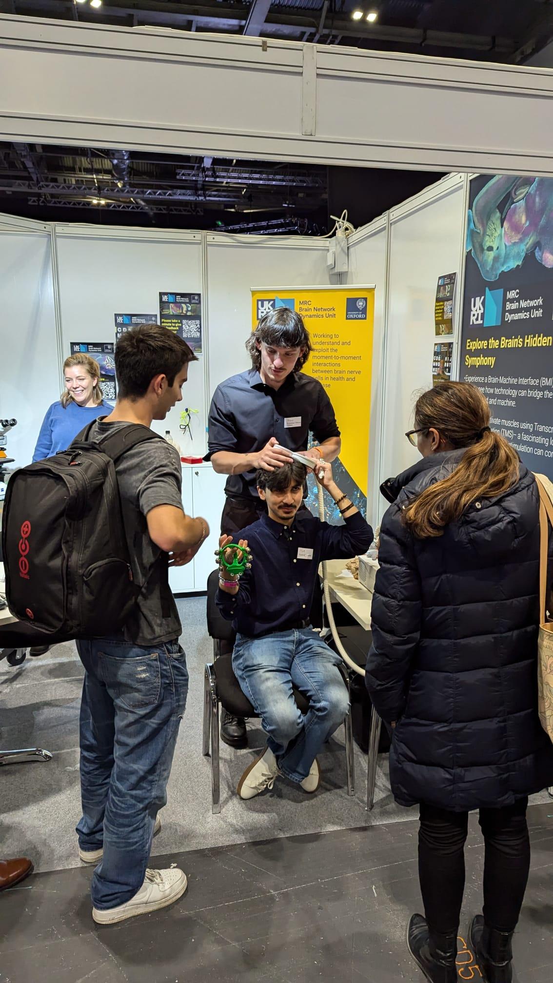 photo of someone holding a magnetic stimulation wand connected via a thick cable, close to another person's head; with people standing around watching