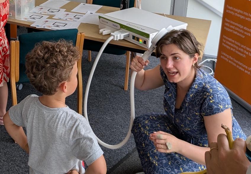 the back of a child, listening to a explanation of transcranial magnetic stimulation, while the scientist holds the stimulation coil to her head.