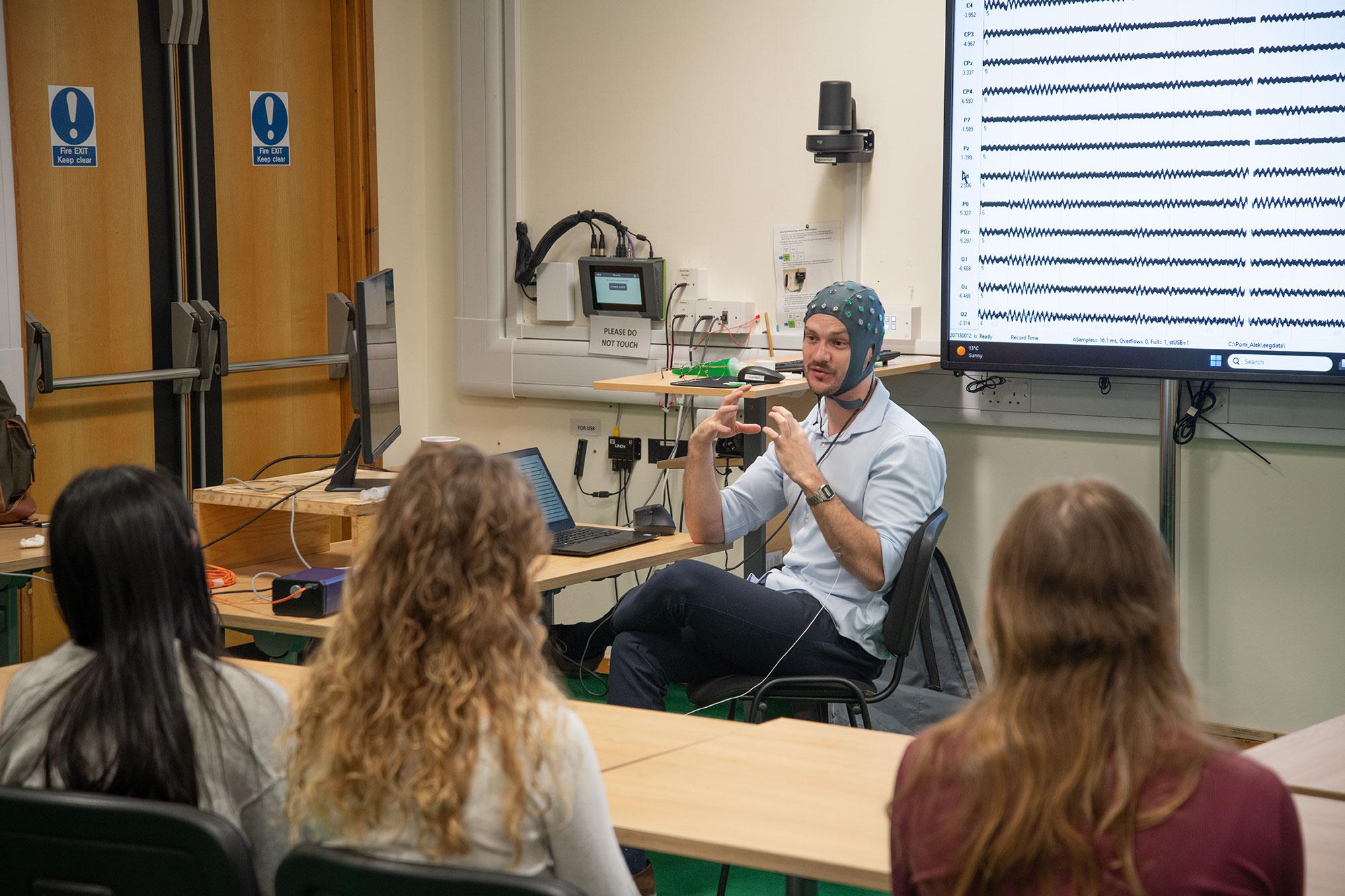 a researcher, wearing an EEG cap, talks to students